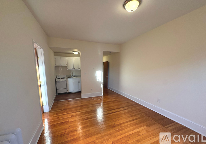 A room with wooden floors and white walls, with a kitchen visible in the background.