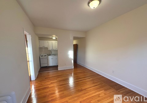 A room with wooden floors and white walls, with a kitchen visible in the background.