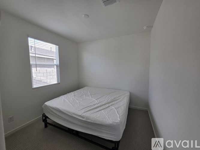 A white bed with a white mattress is in a room with white walls and a window with white blinds.