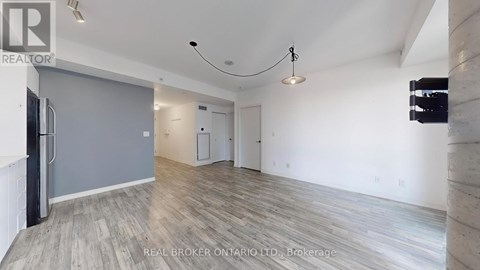 A spacious room with wooden flooring and white walls, furnished with a grey sofa and a black refrigerator.