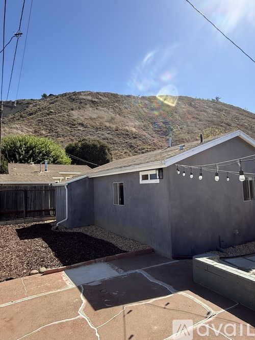 A house with a grey exterior is surrounded by a gravel area and a hill in the background.