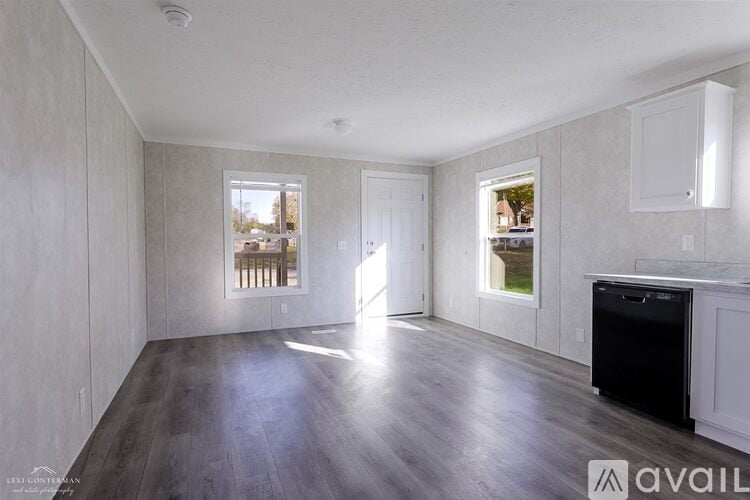 A spacious room with wooden floors and white walls, featuring a large window with a view of trees and a door leading to another room.