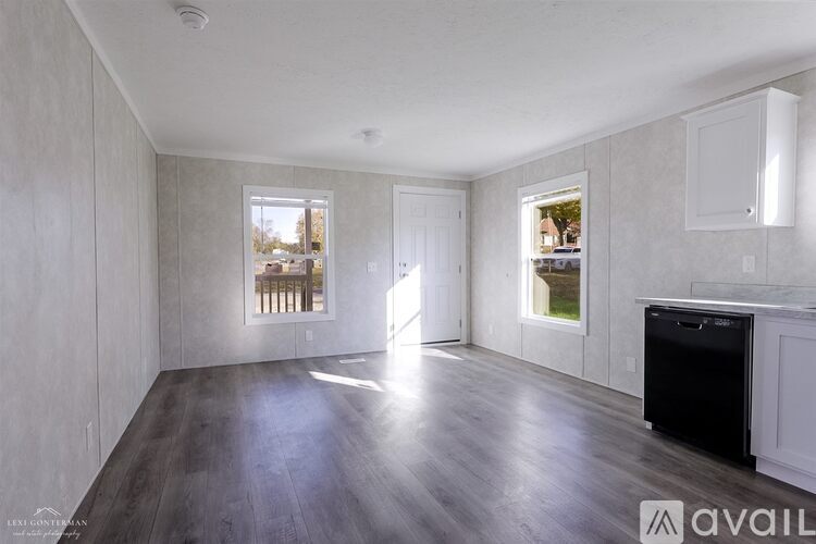 A spacious room with wooden floors and white walls, featuring a large window with a view of trees and a door leading to another room.