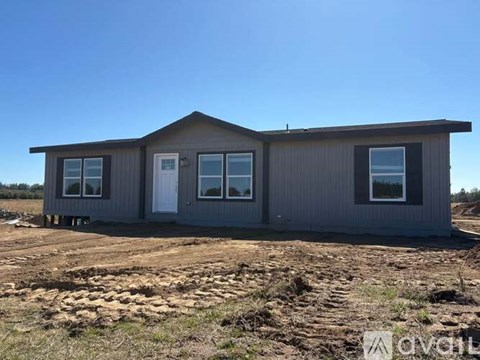 A grey house with a white door and windows is situated on a dirt lot.