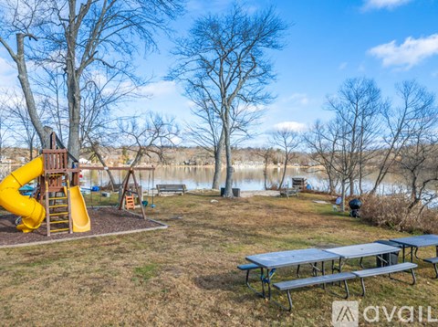 A playground with a yellow slide and picnic tables is surrounded by trees and a body of water.