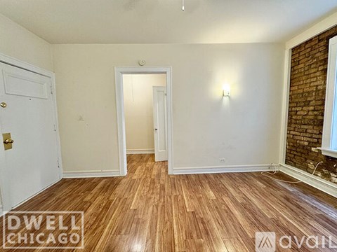 A room with wooden flooring and white walls, with a door on the left and a window on the right.