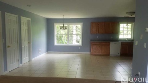 A room with a white door, a window with a view of trees, and a kitchen area with wooden cabinets.