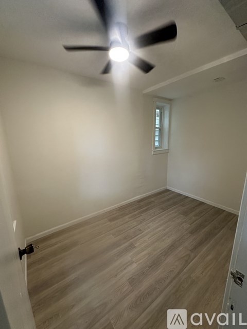 A room with a ceiling fan and light, wooden flooring, and a window with blinds.