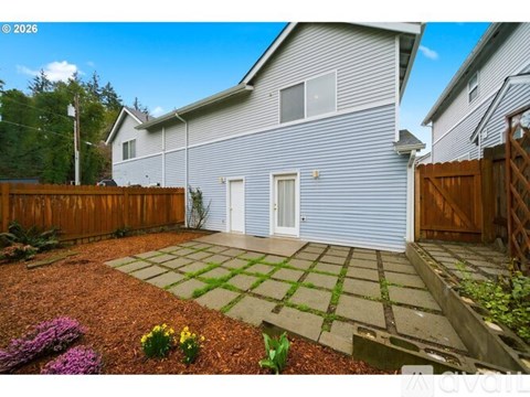 A house with a grey exterior and a white door is surrounded by a wooden fence and a garden with a mix of gravel and grass.