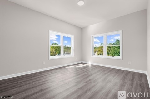 A spacious room with wooden flooring and two windows showing a view of greenery outside.