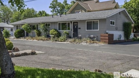 A house with a grey roof and a black door is surrounded by a gravel driveway and a grassy area.