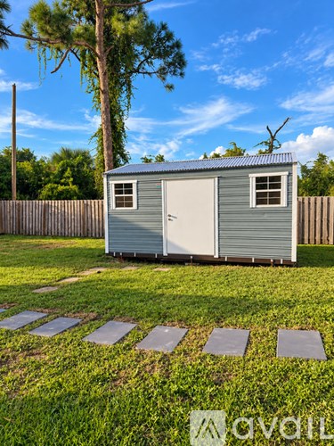 A grey metal shed with a closed grey door and a small window is situated in a grassy area with a wooden fence and a tree in the background.