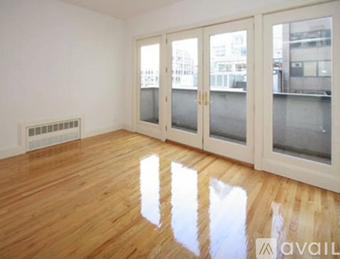 A room with wooden floors and white walls has a window with a view of buildings outside.