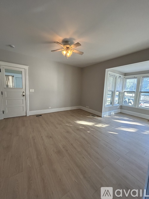 A room with a ceiling fan and light, wooden flooring, and a door leading to another room.