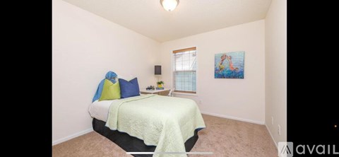 A bedroom with a bed, pillows, a blanket, a window with blinds, a nightstand with a lamp, and a painting on the wall.