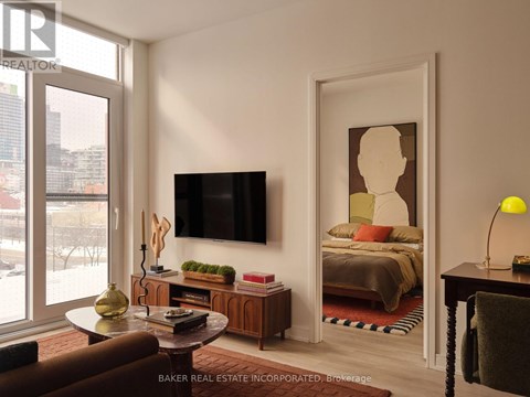 A living room with a brown couch, a flat screen TV, a coffee table, and a bedroom visible through the open door.