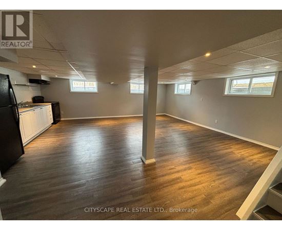 A spacious room with wooden flooring and white walls, with a black refrigerator on the left side.