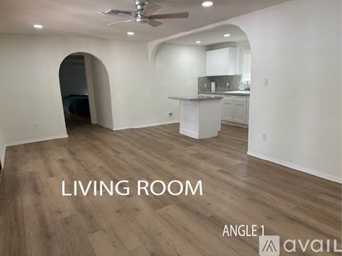 A living room with a kitchen in the background and a ceiling fan.