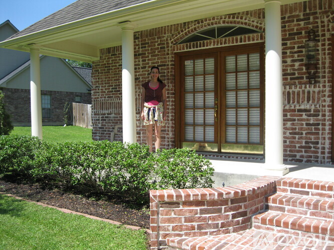 A woman stands on the porch of a brick house.