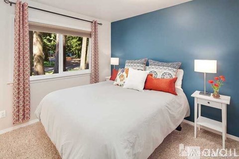 A bedroom with a white bed, a small white nightstand with a lamp and a vase of flowers, and a window with curtains.