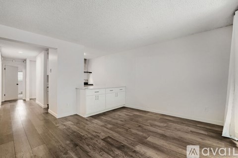 A room with wooden flooring and white walls, with a kitchenette and a doorway leading to another room.