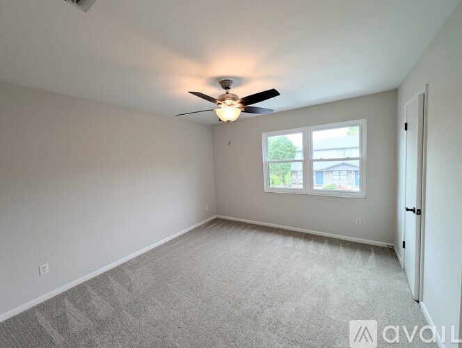 A room with a carpeted floor, a ceiling fan, and a window with a view of trees and a building outside.