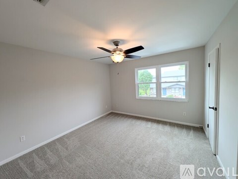 A room with a carpeted floor, a ceiling fan, and a window with a view of trees and a building outside.