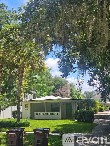 A house with a green door is surrounded by trees and has two trash cans in front.