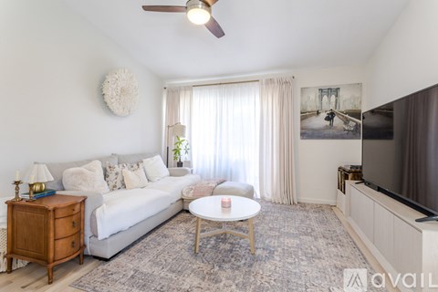 A living room with a white couch, a wooden side table, a white coffee table, a grey rug, a ceiling fan, a painting on the wall, a window with curtains, and a flat screen TV.