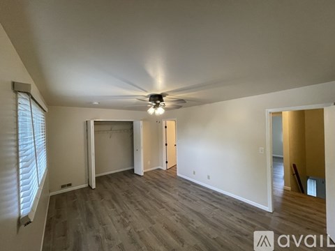 A spacious room with a ceiling fan and light, hardwood flooring, and a large window with blinds.