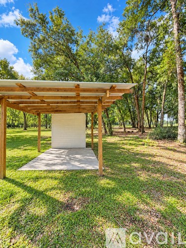 A pavilion-like structure with a white roof and wooden pillars is situated in a grassy area with trees in the background.