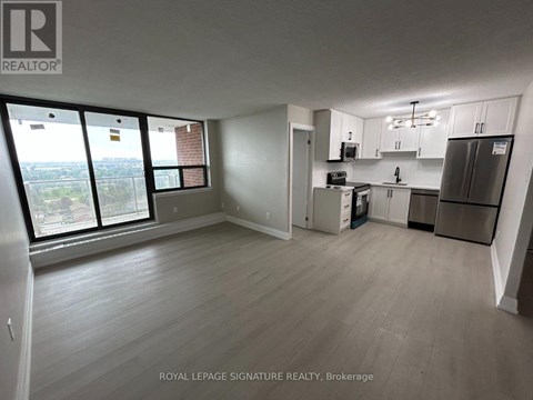 A well-lit, empty room with a kitchenette and a view of the outside through large windows.