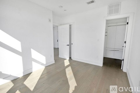 A spacious room with white walls and wooden flooring, featuring a large window on the left and a door on the right.