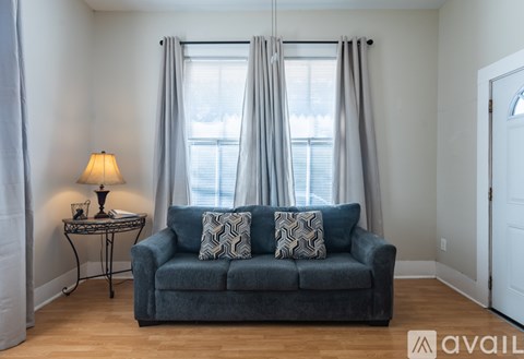 A living room with a grey couch, a small table with a lamp, and a window with curtains.