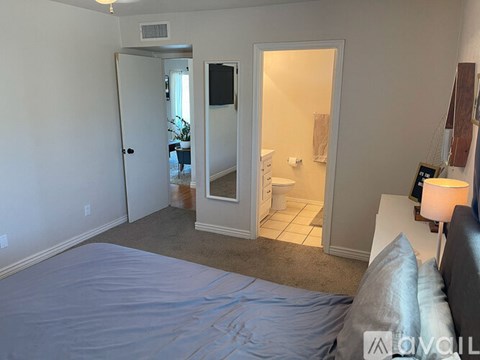 A bedroom with a bed covered in a blue sheet, a lamp on a side table, and a bathroom visible through a doorway.