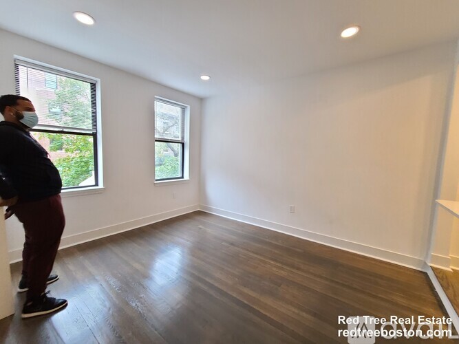 A man wearing a mask stands in a room with wooden floors and white walls.