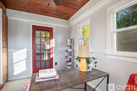 A room with a red door, a wooden table with a lamp and a plant on it, and a window with a view of trees outside.
