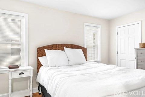 A bedroom with a white bed, a small table, and a window with blinds.