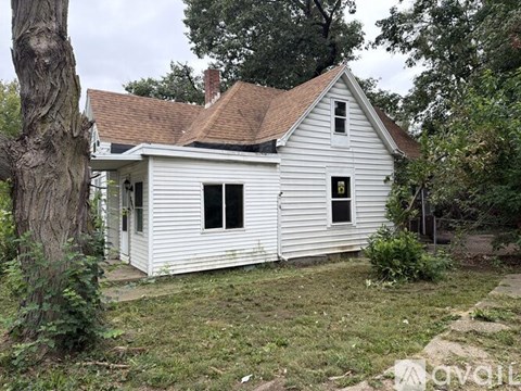 A small white house with a brown roof and a chimney is surrounded by trees and grass.