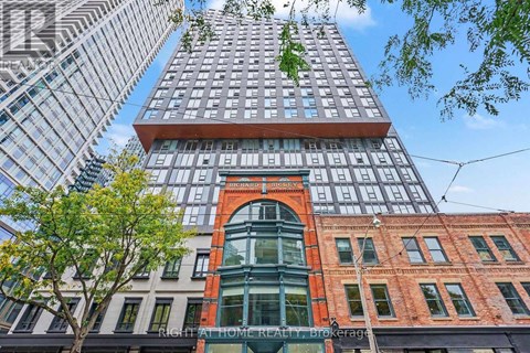A tall building with a glass facade stands in front of a tree and other buildings.