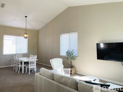 A living room with a grey couch, a white chair, a dining table with chairs, a television, and a potted plant.