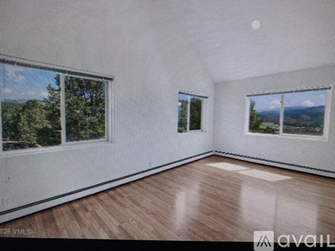 A room with wooden flooring and three windows showing a view of trees and sky.