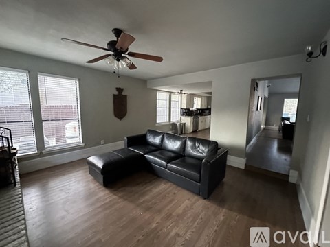 A black leather couch sits in a spacious living room with a ceiling fan and large windows.