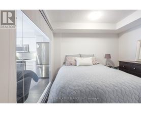A bedroom with a large bed and a window overlooking a kitchen.