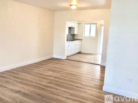 A room with wooden flooring and white walls, with a kitchen area visible in the background.
