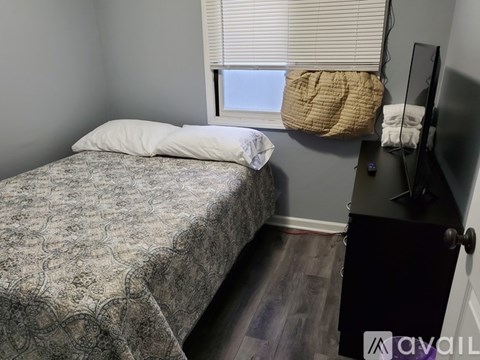 A bedroom with a bed, a window with blinds, a TV on a stand, and a wall-mounted shelf.