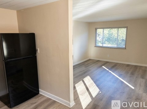 A black fridge sits in a room with wooden floors and a window.