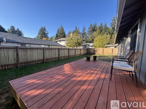 A wooden deck with a chair and a table is in the foreground with a house and trees in the background.