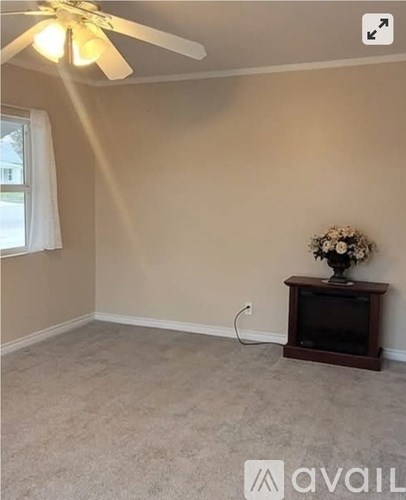 A room with a ceiling fan, a window with white curtains, and a wooden entertainment center with a vase of flowers on top.