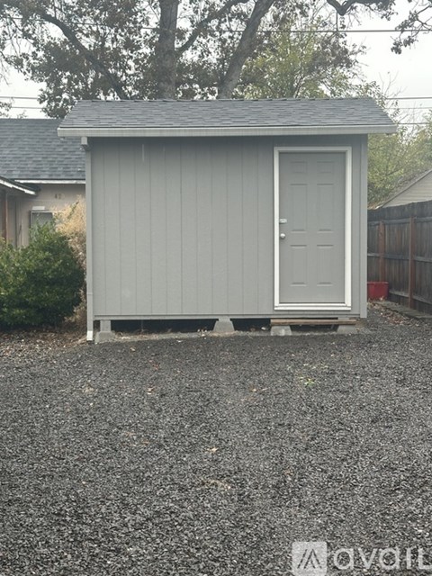 A small grey shed with a white door is situated in a gravel area.